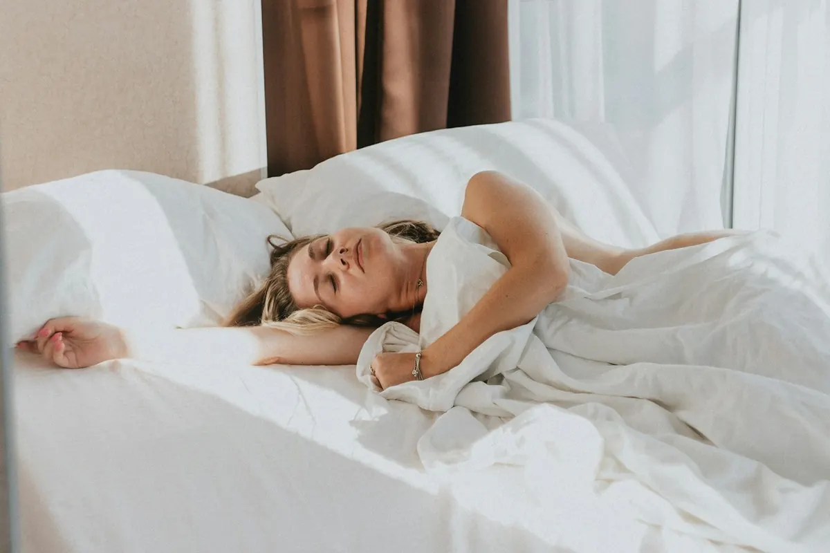 Femme allongée dans un lit aux draps blancs, les yeux fermés, un bras étendu, dans la lumière du matin