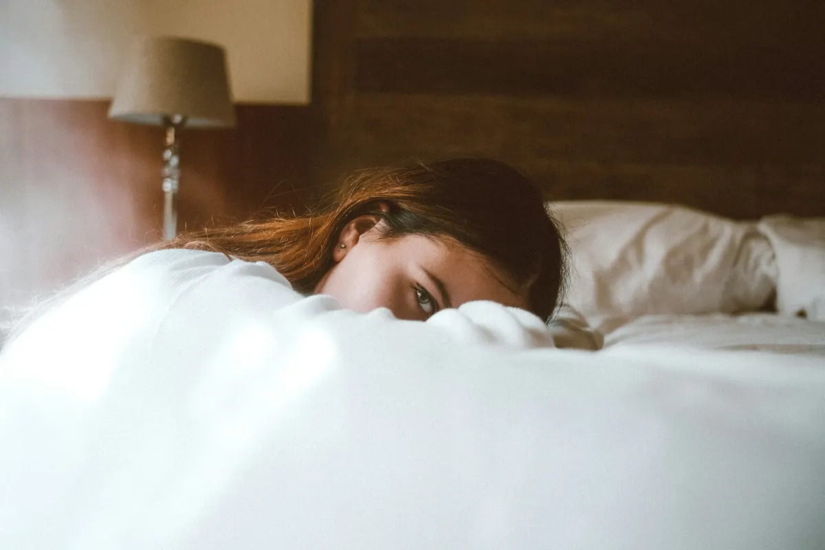 Femme allongée sur son lit, le visage à moitié caché par la couette, les yeux ouverts, aux aguets