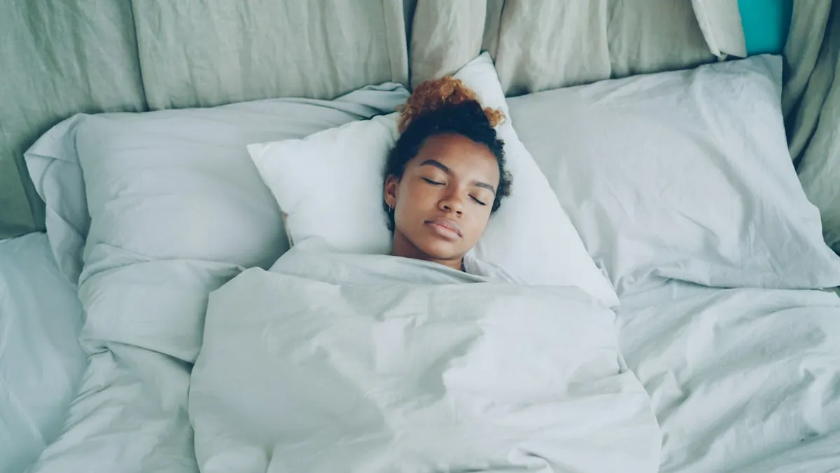 Jeune femme allongée sur le dos dans un lit blanc, les yeux fermés, le visage détendu, la couette remontée jusqu'aux épaules