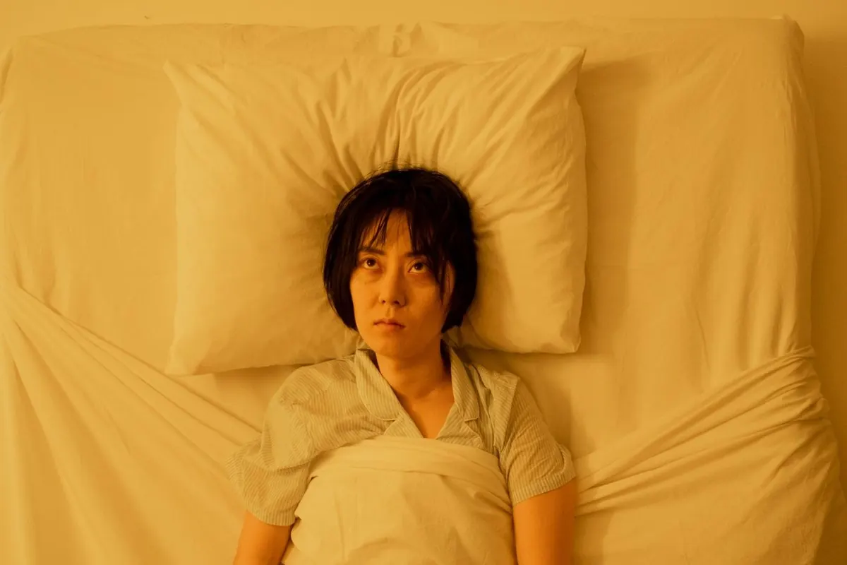 Femme allongée sur le dos dans un lit aux draps clairs, vue d'en haut, les yeux grands ouverts et les bras le long du corps, dans une lumière chaude